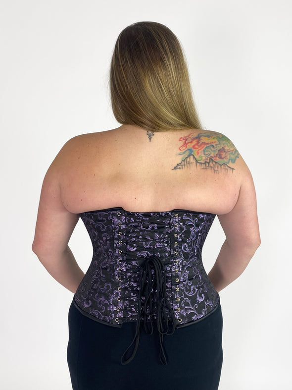 back lace up corset view of a plus size model wearing a purple brocade overbust steel boned corset top with black leggings